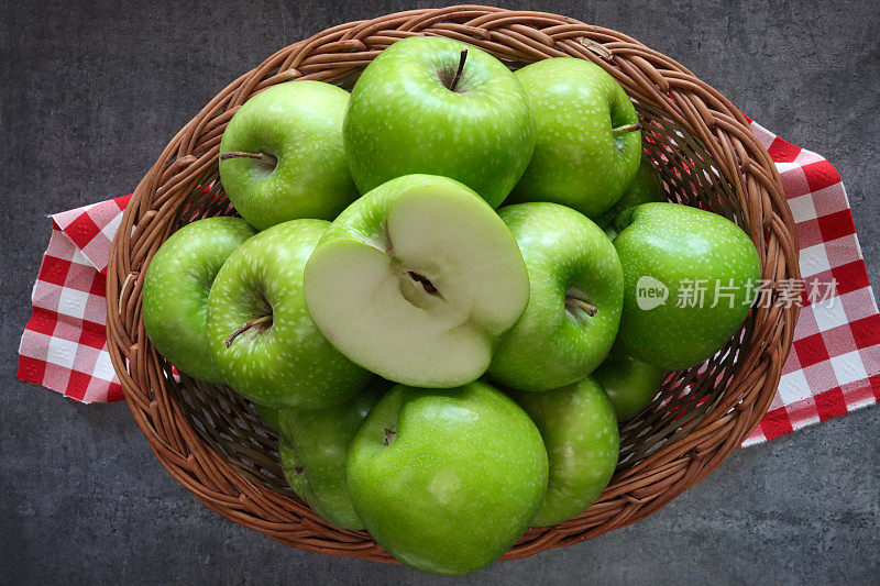 红白格子布上的柳条篮子的特写图像，在一堆绿色的史密斯苹果(Malus domestica)上切了一半的水果，有茎，光滑的，有斑点的果皮，斑驳的深灰色背景，高架视图